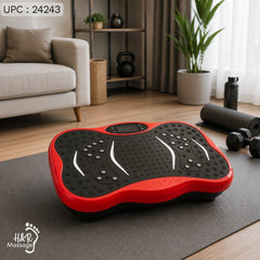 A red and black vibration plate exercise platform placed on a dark yoga mat in a modern living room setting with a sofa, plants, and dumbbells visible in the background.